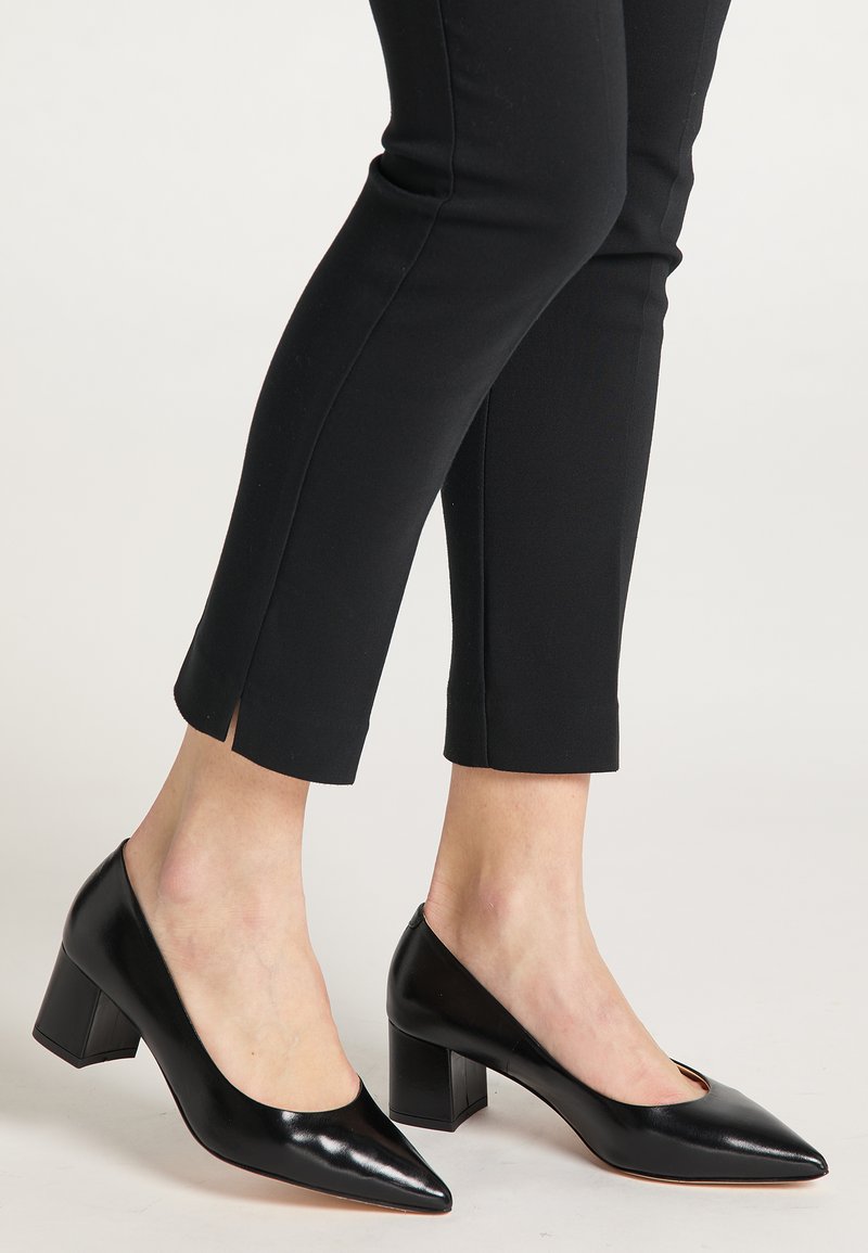 Zwarte leren pumps met hoge hakken en een puntige neus, voorzien van een blokhak en zij-snijdetails, gecombineerd met zwarte, taps toelopende broek.