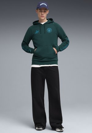 Junger Mann, der einen dunkelgrünen Manchester City Hoodie, schwarze weit geschnittene Hosen, Turnschuhe und eine dunkelblaue Baseballkappe mit der Aufschrift "MAN CITY" trägt, steht mit den Händen in den Taschen.