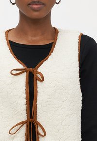 Fuskepelsvest i kremhvit med brune suedekanter, med knyting foran og teksturert overflate. Brukt over en svart topp.