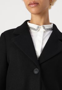 Cappotto di lana nero con colletto a punte, dotato di chiusura con bottoni. Sotto, una camicia bianca con dettagli del colletto gioiello.