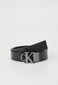 Črn usnjen pas z pravokotno srebrno sponko, ki nosi inicialke "CK." Pas je gladek z rahlim leskom in se zoži proti koncu.