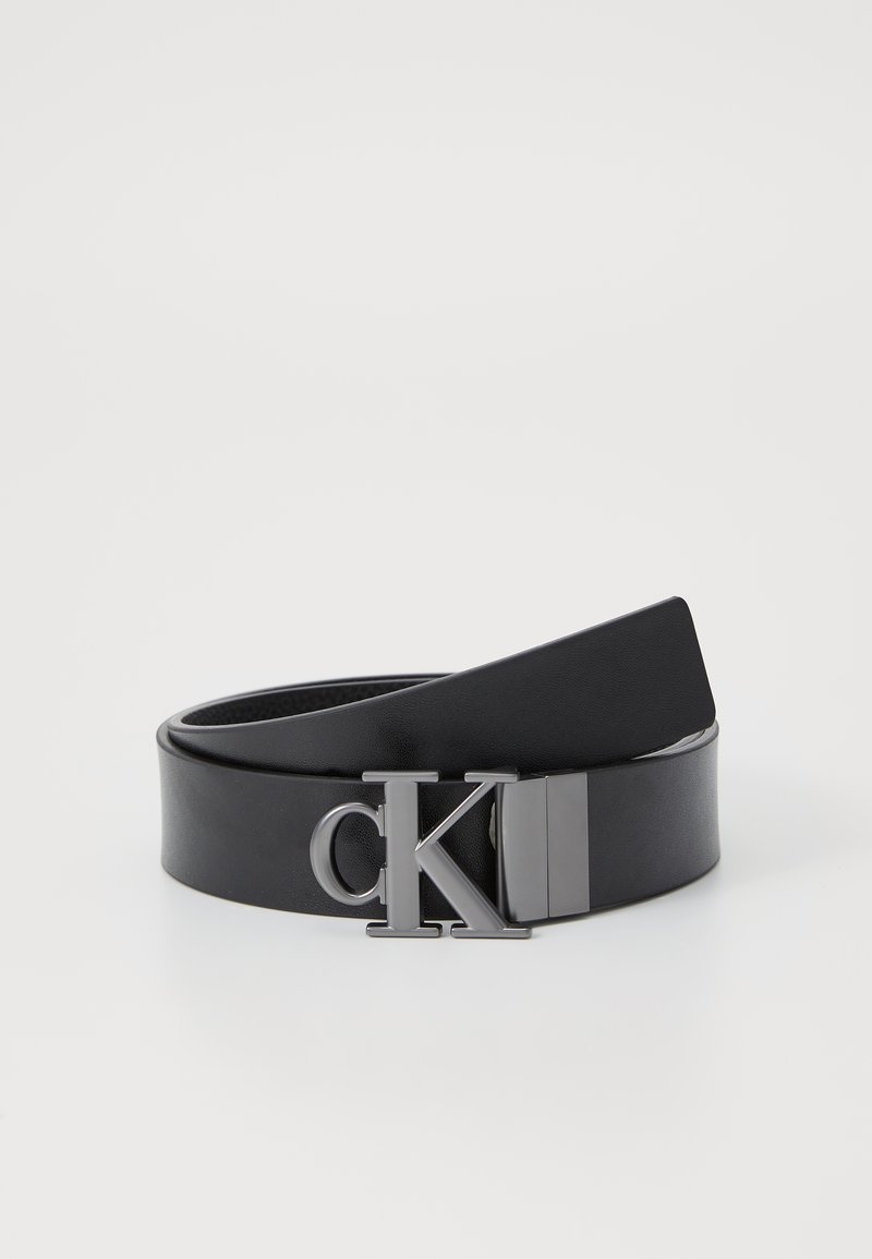 Svart lærbelte med en rektangulær sølvspenne som har initialene "CK." Remmen er glatt med en svak glans og avsmalner mot enden.
