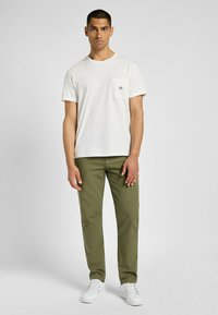 Biała bawełniana koszulka z kieszonką na piersi, zestawiona z oliwkowymi spodniami i białymi trampkami, stojąca na jednolitym tle.