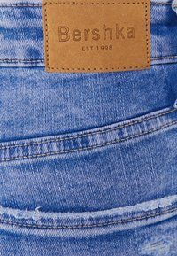 Lichtblauwe spijkerbroek van denim met een gestructureerde leren patch waarop "Bershka" en "EST.1998" is geperst, met zichtbare stiksels en een versleten zoom.