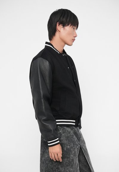 Giacca varsity nera con corpo in lana, maniche in pelle, colletto a coste con strisce e chiusura a bottoni a pressione. Indossata con pantaloni grigi a fantasia.