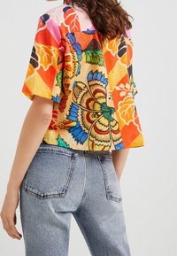 Kortærmet, cropped top i farverige blomster- og geometriske mønstre, lavet af let stof, parret med mellemhøje blå jeans.