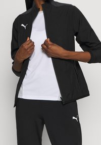 Zwart lichtgewicht jack met een hoge kraag en wit Puma-logo, gecombineerd met een wit shirt en zwarte broek. Glad materiaal met een aansluitend ontwerp.