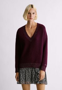 Maglione bordeaux a maglia con scollatura a V, tessuto strutturato e orlo con dettagli metallici, abbinato a una gonna con motivo in bianco e nero.