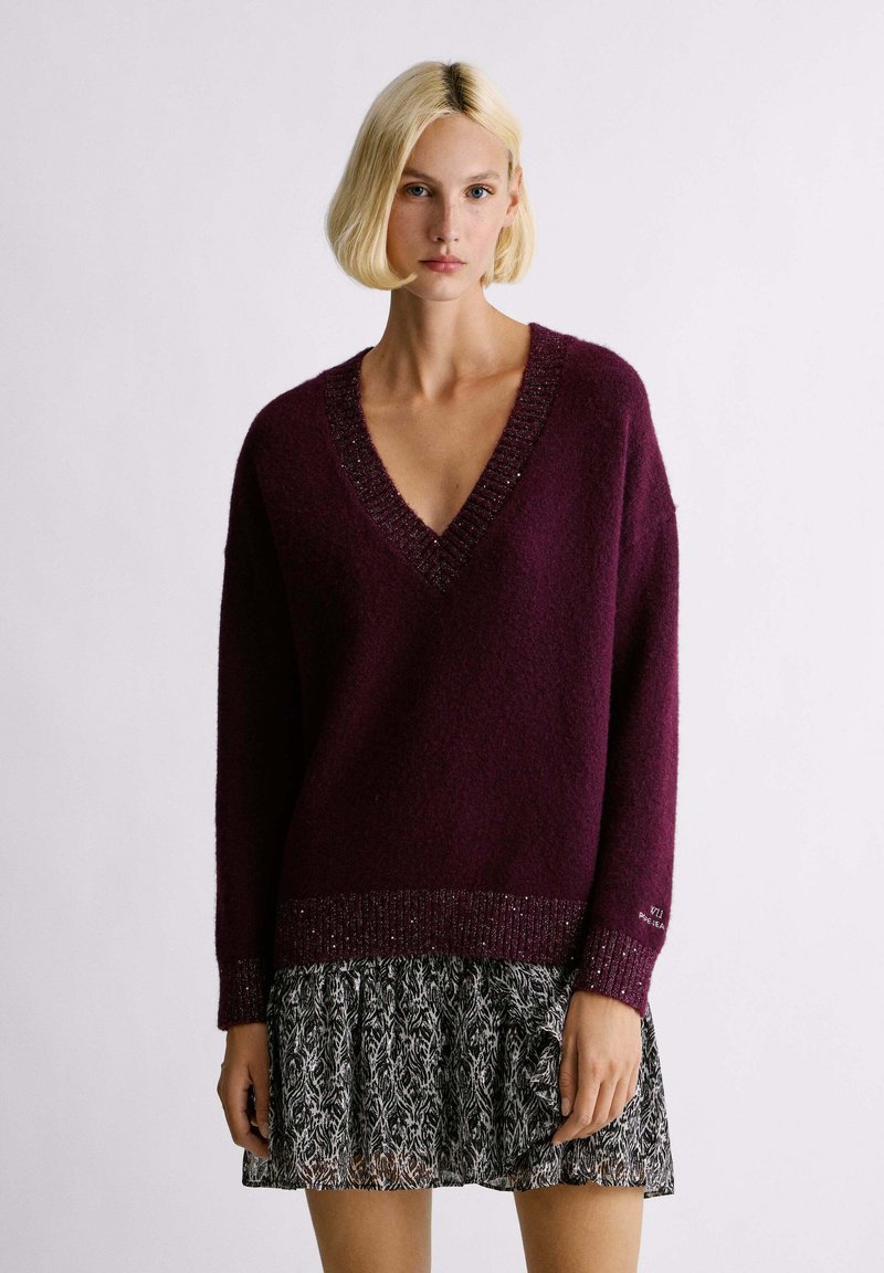 Maglione bordeaux a maglia con scollatura a V, tessuto strutturato e orlo con dettagli metallici, abbinato a una gonna con motivo in bianco e nero.