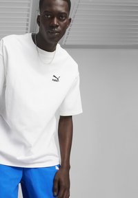 Hvid bomulds t-shirt med et sort Puma-logo på brystet, båret med blå shorts. T-shirten har en afslappet pasform og korte ærmer.