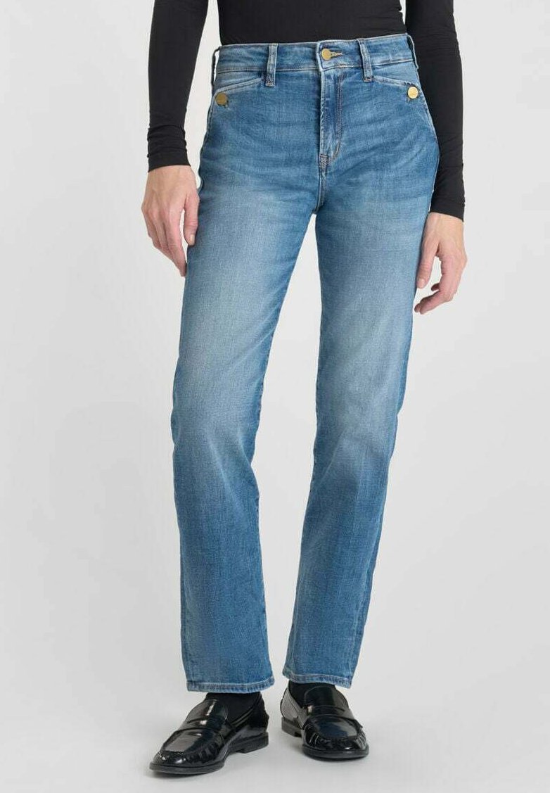 Jean en denim bleu clair avec une coupe droite, taille haute, accents de boutons dorés et léger délavage. Porté avec des mocassins en cuir verni noirs.