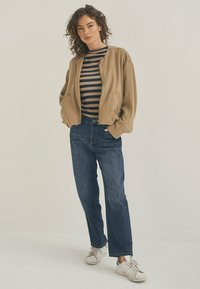 Khaki Bomberjacke über einem schwarz-weiß gestreiften Hemd, kombiniert mit blauen geraden Jeans und weißen Turnschuhen.