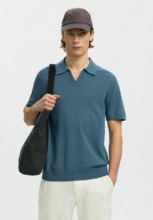 Junger Mann, der ein blaues Kurzarm-Poloshirt, weiße Hose, graue Kappe trägt und eine schwarze Umhängetasche bei sich hat, steht vor einem schlichten Hintergrund.