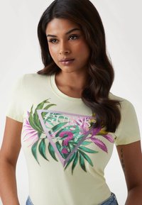 T-shirt en coton jaune clair avec un graphique triangulaire coloré représentant des feuilles tropicales et du texte de logo, manches courtes et un design ajusté.