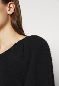 Orecchio sinistro di una donna con un piccolo orecchino a cerchio dorato, lunghi capelli castani, che indossa un maglione nero e peloso su uno sfondo chiaro e semplice.