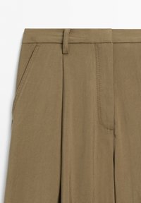Close-up of brown pants waist with belt loops, front pleat, and side pocket stitching on a white background.