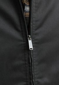 Gros plan d'une veste noire partiellement zippée avec un tirette de fermeture éclair argentée montrant le nom de la marque "Jack & Jones" et une chemise à carreaux à l'intérieur.