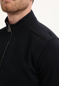 Primer plano de un hombre que lleva una chaqueta negra con cremallera hecha de un tejido texturizado y un tirador de cremallera metálico con un diseño geométrico.
