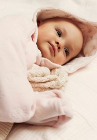 Baby gewikkeld in een lichtroze, gestructureerde deken met een bloemenpatroon kap, rustend op een zacht, neutraal, gewatteerd oppervlak.