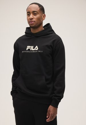 Hombre con sudadera negra con capucha con logo blanco de FILA y texto "Established 1911," de pie con las manos en los bolsillos contra un fondo claro y liso.