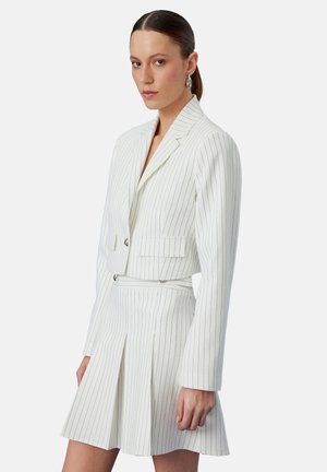 Kvinde iført hvid blazer med nålestriber og matchende plisseret nederdel, med håret trukket tilbage og sølvfarvede creoler, mod ensfarvet baggrund.