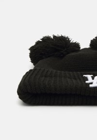 Fekete kötött sapka, két pom-pommal a tetején, bordázott szegéllyel és fehér hímzett részlettel az elején. Puha tapintású és kerek formájú.