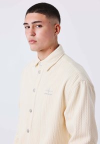 Camisa de botón color crema con un patrón de punto texturizado, que presenta botones plateados y un pequeño logo en el pecho.