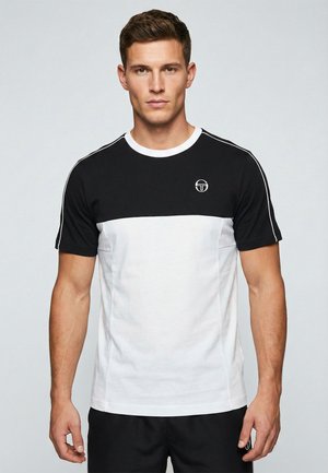 Hombre con una camiseta de manga corta en blanco y negro con un logo en el pecho, de pie frente a un fondo liso y claro.