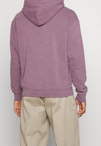 Felpa di cotone viola con tasca a marsupio, cuciture frontali e polsini a costine, abbinata a pantaloni beige. Design morbido e informale.