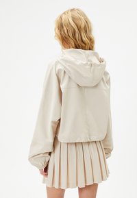 Chaqueta corta beige con capucha, con puños elásticos y una textura suave, combinada con una falda plisada en un color a juego.