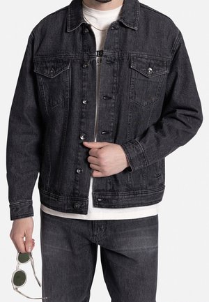 Mężczyzna ubrany w czarną kurtkę denim i jeansy na białej koszulce, trzymający okulary przeciwsłoneczne z przezroczystą oprawką w lewej ręce na białym tle.