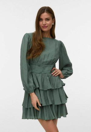 Naine kannab salvei-rohelist tekstuuriga kleiti, millel on pikad pallimustrilised varrukad ja kihiline volangiline seelik, poseerides ühe käega vööl.