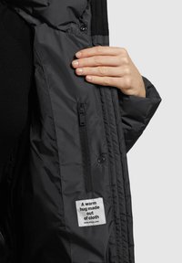 Een hand houdt een open zwarte jaszak vast, waar een rits en een label zichtbaar zijn met de tekst "Een warme omhelzing van stof" en een website-link.