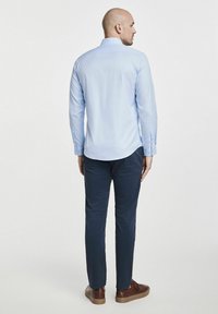 Jasnoniebieska koszula z długim rękawem i zapiętymi mankietami, w połączeniu z ciemnoniebieskimi spodniami i brązowymi butami, widziana z tyłu na neutralnym tle.