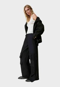 Pantaloni cargo neri con multiple tasche, abbinati a una canotta bianca aderente e a una giacca nera ampia, con scarpe flat nere a pois.