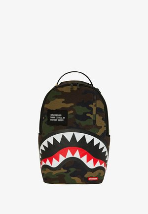 Mochila camuflada con un gráfico de boca de tiburón negra, detalles en rojo y dientes blancos; las etiquetas de tela dicen "Sprayground" y exhiben detalles de ubicación.