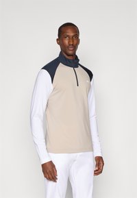 Μπεζ μισό φερμουάρ μπλούζα μακρύ μανίκι με σκούρες μπλε λεπτομέρειες στους ώμους, φτιαγμένη από απαλό ύφασμα. Διαθέτει φερμουάρ στον λαιμό και λευκά μέρη στα μανίκια.