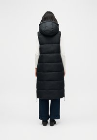 Persona con un chaleco largo acolchado negro con capucha, camiseta blanca de manga larga, pantalones anchos oscuros y zapatos negros de tacón, de pie de espaldas.