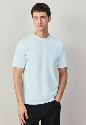 Fiatal férfi sima világoskék rövid ujjú pólóban és fekete nadrágban, egyik kezét zsebében tartva, világos háttér előtt áll.