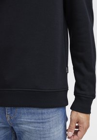 Felpa nera con orlo e polsini a costine, dotata di un piccolo etichetta del marchio sul lato. Indossata con jeans blu. Tessuto morbido.