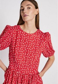 Rotes Blumenkleid mit kurzen Puffärmeln, rundem Ausschnitt und gerafftem Taillenbereich. Ausgestattet mit einem weißen Blumenmuster auf einem strukturierten Stoff.
