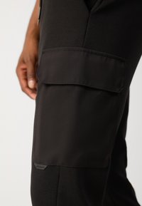 Svarte cargo bukser har en stor sidelomme med klaff, laget av et glatt, elastisk stoff. Teksturert detaljering nær lommen fremhever designet.