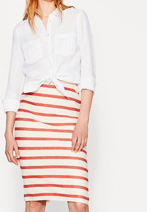 Wit linnen overhemd met opgerolde mouwen en knoopsluiting aan de voorkant, geknoopt in de taille over een gestreepte rok tot op de knie in rood en crème.