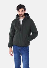 Giacca verde imbottita con cappuccio, dotata di zip frontale e polsini elastici. Abbinata a jeans di denim blu, esposta su uno sfondo neutro.