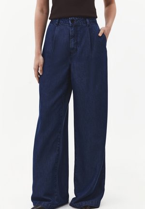 Kvinde iført mørkeblå jeans med høj talje og vide ben, med forreste læg, sidelommer og en sort ærmeløs top.
