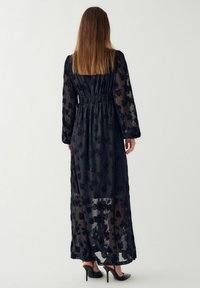 Czarna długa sukienka z długim rękawem, przezroczysta tkanina z florystycznym wzorem, marszczona talia, do ziemi, zestawiona z czarnymi szpilkami.