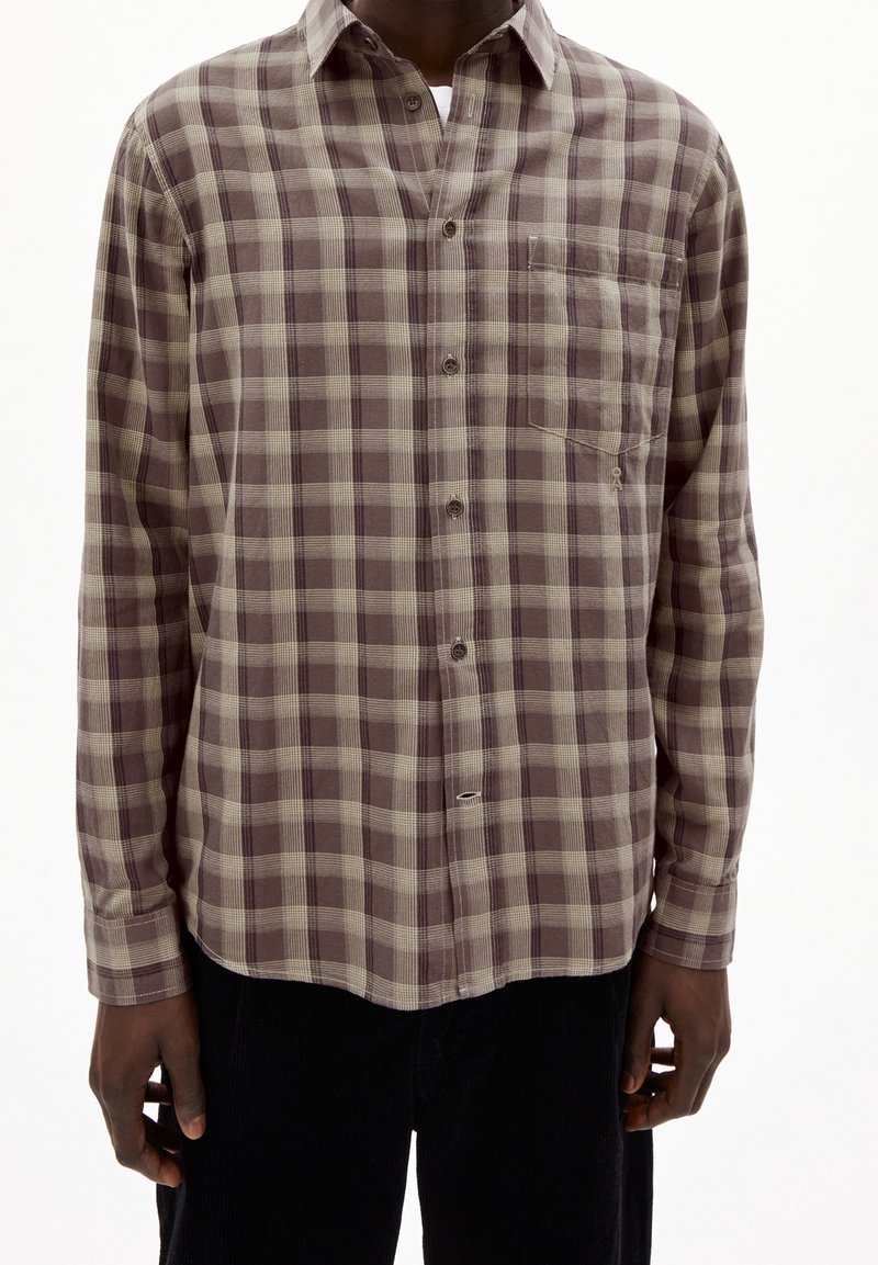 Homme portant une chemise à carreaux marron et beige à manches longues avec une poche poitrine, visible des épaules aux hanches.