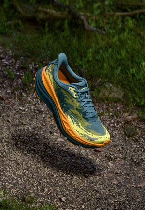Chaussure de course avec une tige principalement verte, des accents jaunes et oranges, un design graphique texturé et une semelle épaisse pour le soutien.