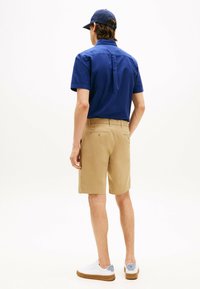 Chemise bleu à manches courtes, shorts kaki, vue de dos. La chemise a un col et un empiècement au dos ; les chaussures sont blanches avec des accents bleus.