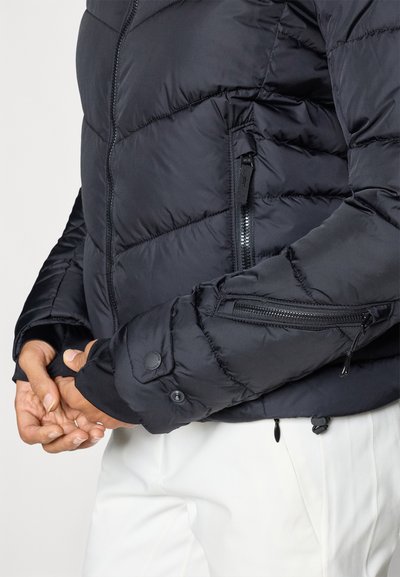 Casaco puff preto com design acolchoado, bolso com fecho éclair na manga e punhos com botões de pressão. Textura suave e material leve.